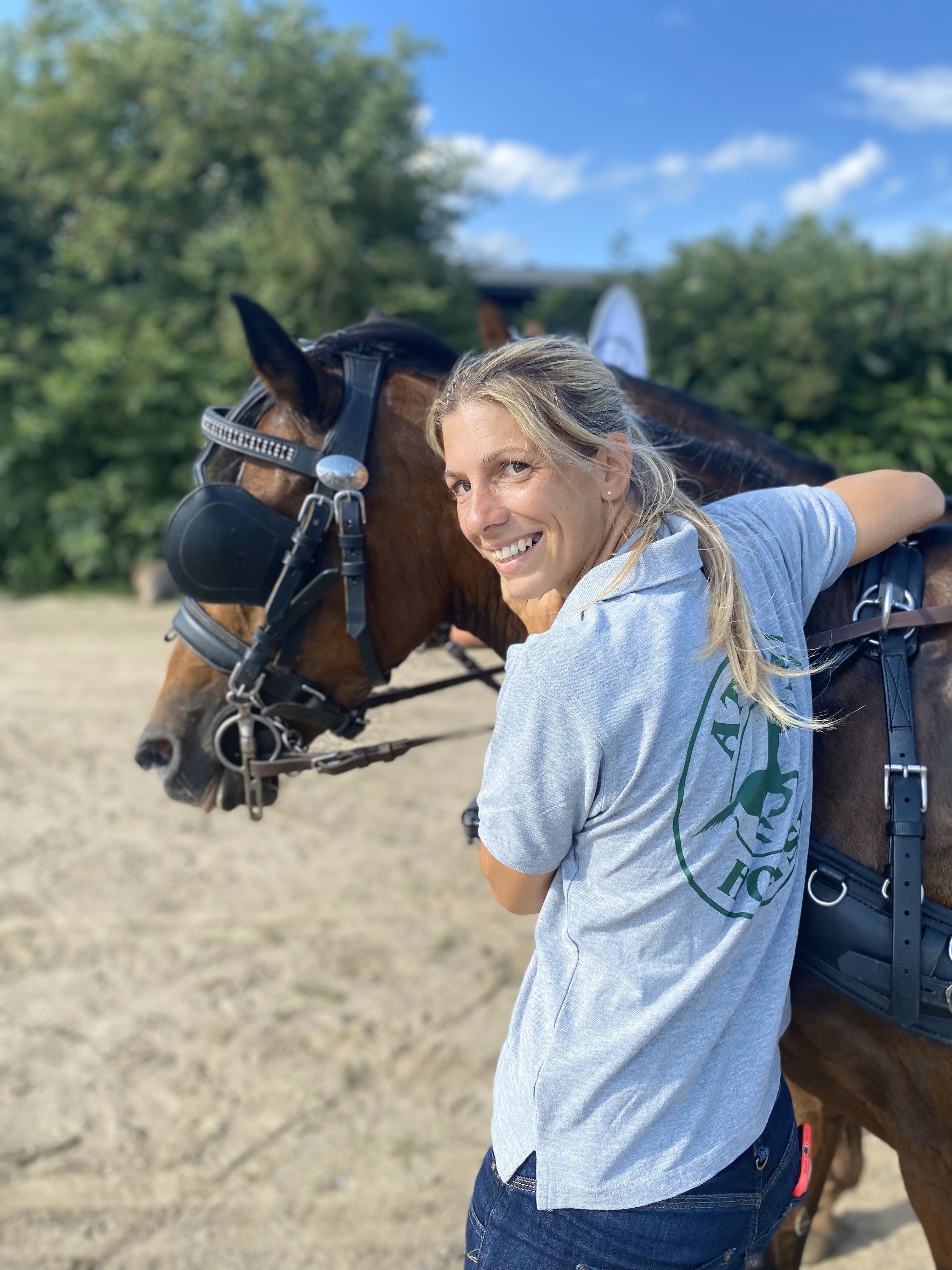 Frau umarmt ein Pferd Eine Frau mit langen blonden Haaren lächelt, während sie ein Pferd mit einer Augenmaske umarmt. Sie trägt ein graues Polo-Shirt mit einem Logo.
